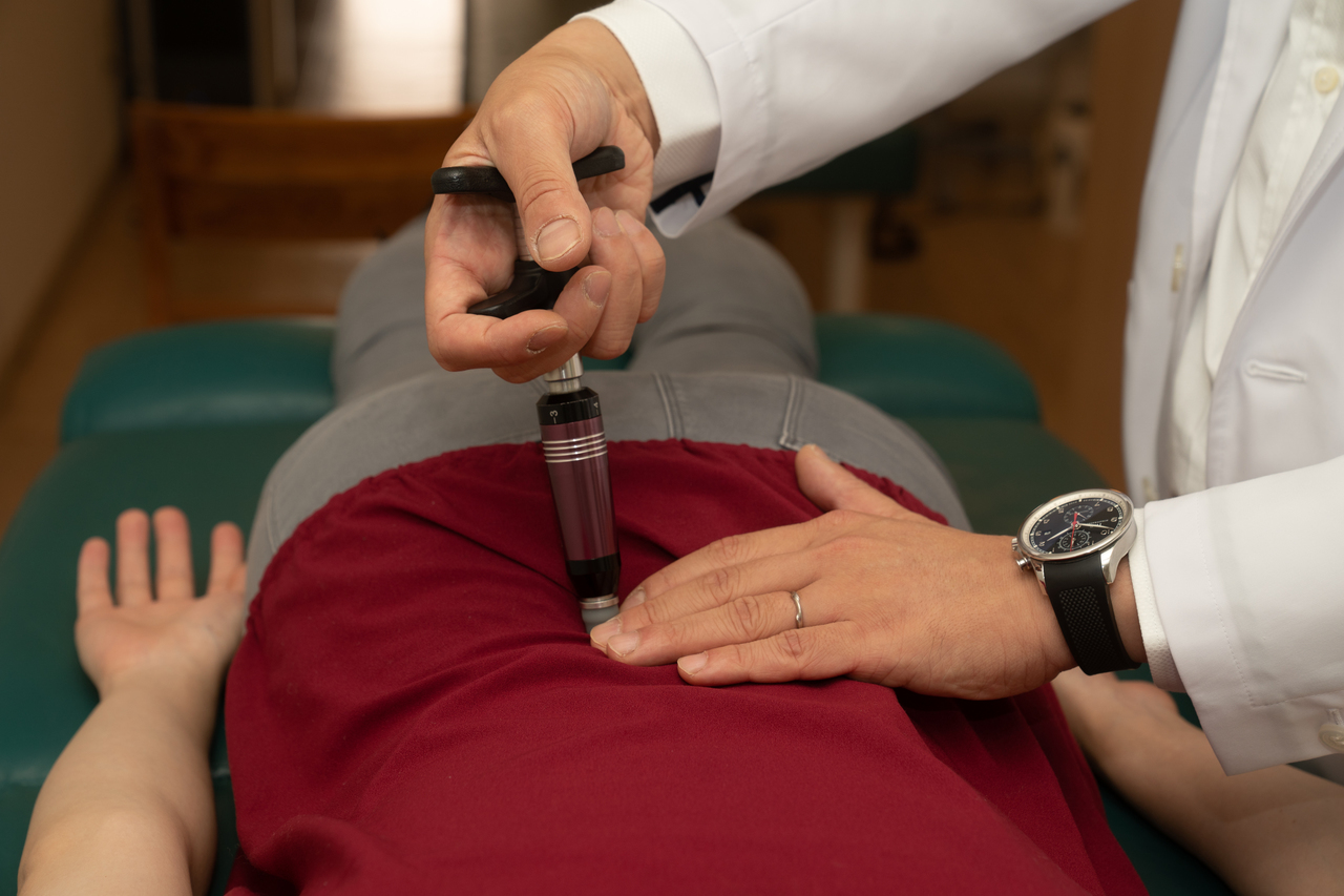 施術ベッドにうつ伏せになった患者に、施術者がアクティベータ器具で背中の調整を行っている様子